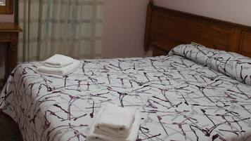 Una cama bien tendida con una colcha estampada y toallas dobladas, un cabecero de madera, una lámpara de pared y una ventana con cortinas.