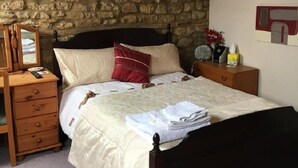 A bedroom with a stone wall, a wooden dresser, a bed with a dark headboard, and a nightstand with a lamp and a clock. - The Lampet Arms (Banbury)