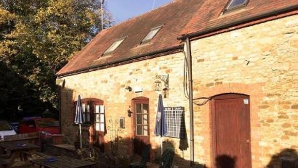 A stone building with a tiled roof, a wooden door, and a small table and chair outside. - The Lampet Arms (Banbury)