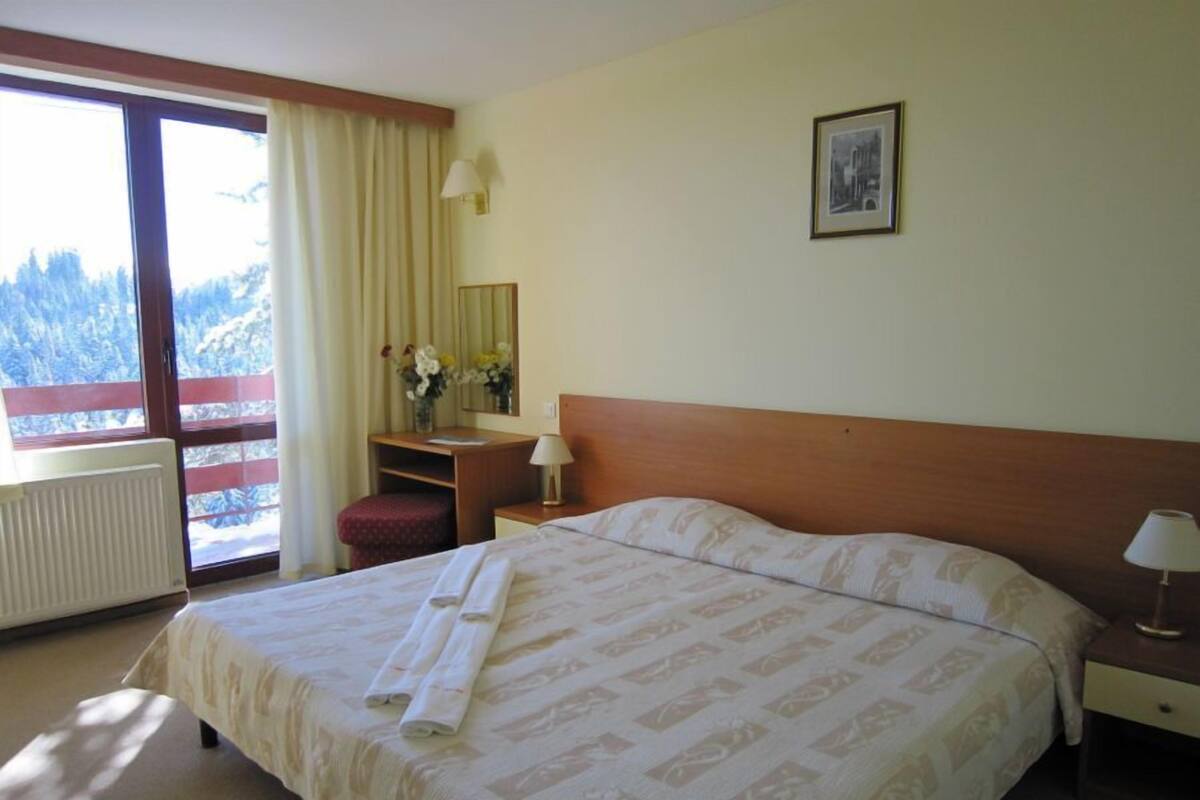 A hotel room with a large bed, a wooden headboard, a nightstand, a desk with a lamp, a window with curtains, and a framed picture on the wall.