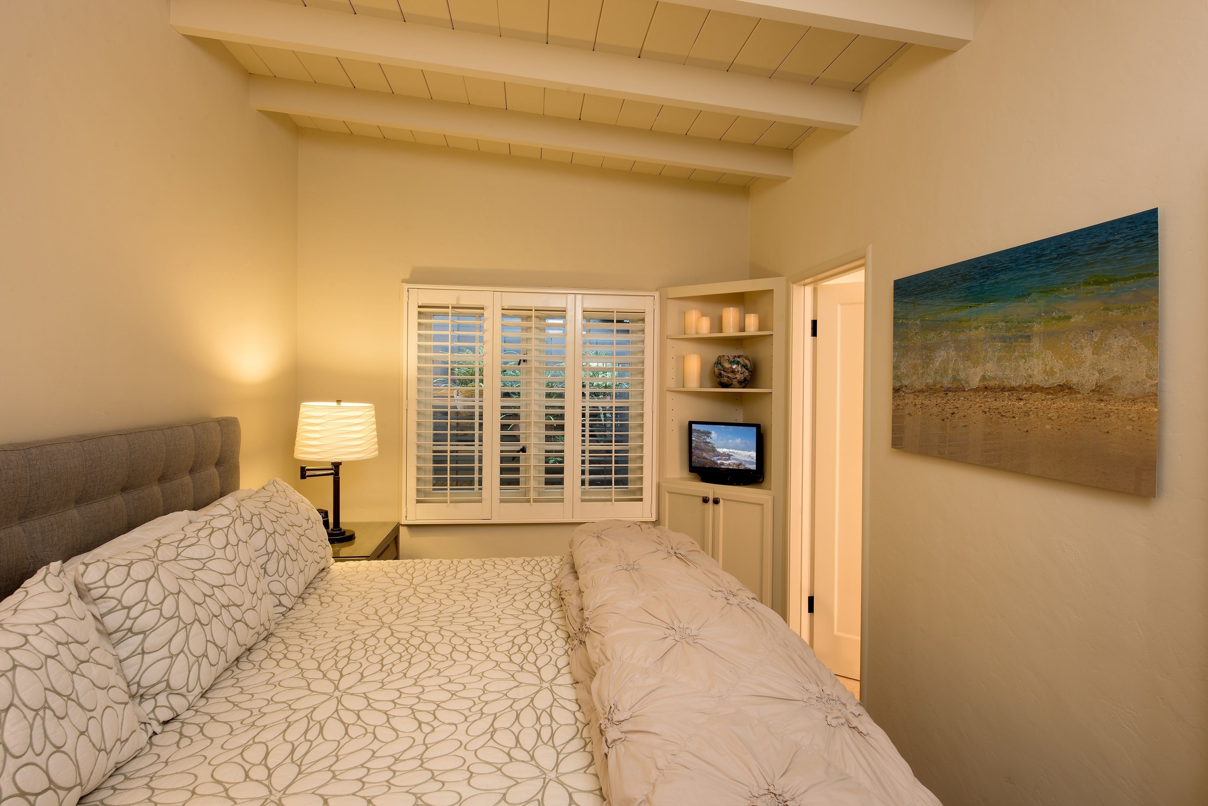 Un dormitorio con cama, ventana con persianas, televisor en un estante y un cuadro en la pared.
