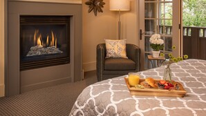 A hotel room with a fireplace, a bed with a tray of breakfast, a chair, a lamp, and a view of the outdoors. - Carmel Country Inn (Carmel)