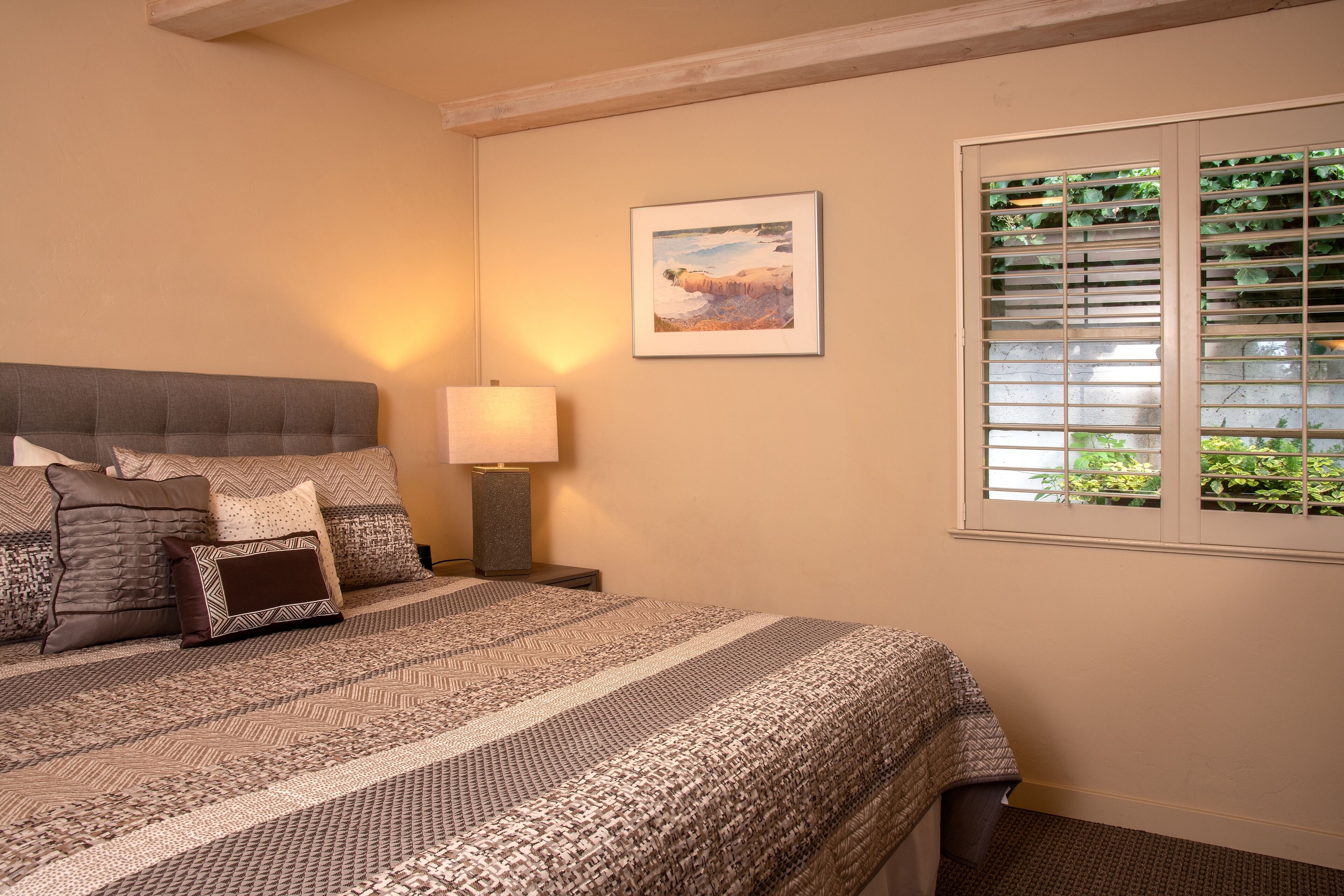 A neatly made bed with patterned bedding, a bedside lamp, and a framed landscape painting on the wall.
