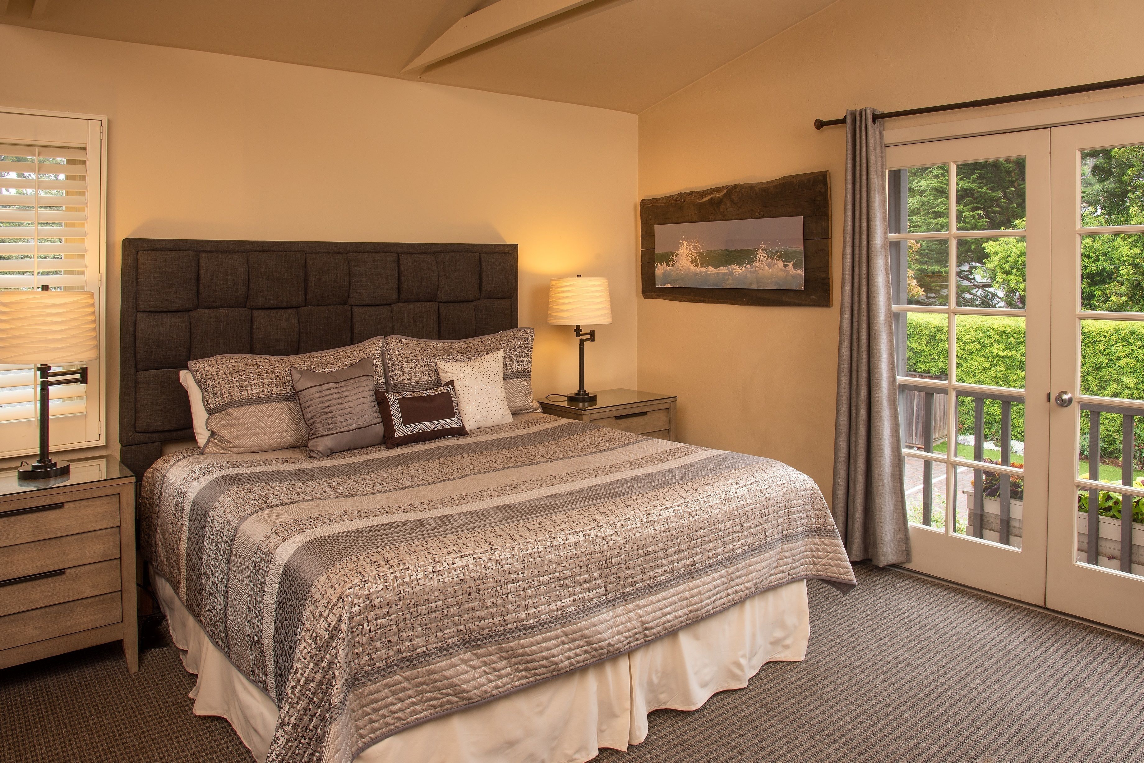A bedroom with a large bed, a nightstand, a lamp, a painting, and a view through a glass door.