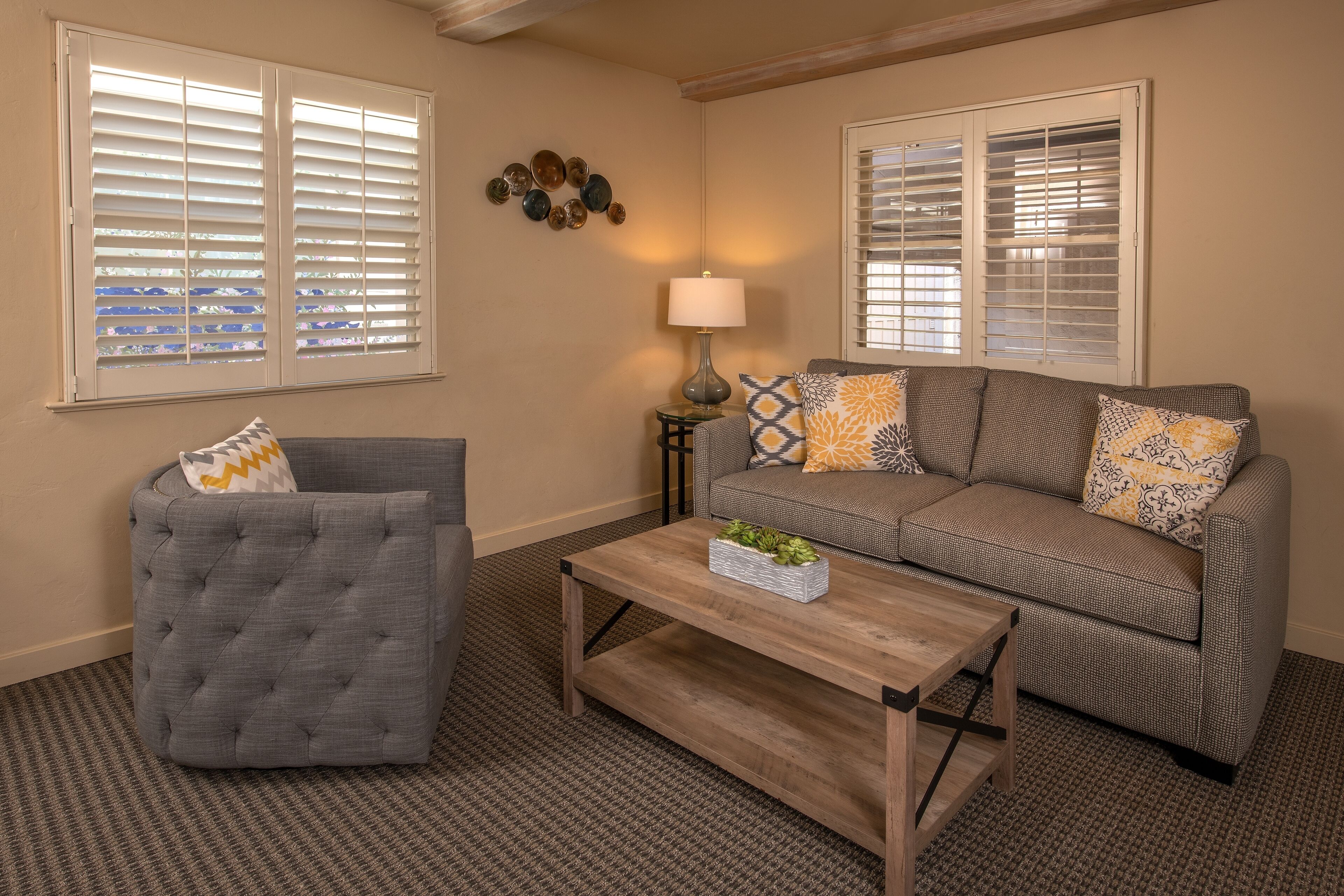 A living room with a grey tufted armchair, a sofa with patterned cushions, and a wooden coffee table.