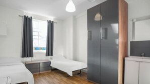 A modern hotel room with two single beds, a large wardrobe, a bathroom with a mirror and sink, and a window with curtains. - Bowden Court - Hostel (London)