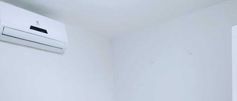A small, clean room with a single bed, a bedside table, and a wall-mounted air conditioning unit.