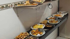 A buffet setup with various food items on trays and baskets, a sign indicating 'Café da Manhã (Breakfast)', and a tiled wall with a decorative border. - Pousada Tambaú (João Pessoa)