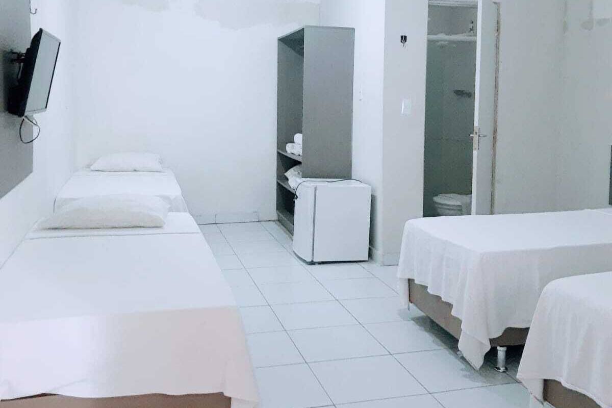 A hotel room with three single beds, a small white refrigerator, a TV mounted on the wall, and a walk-in closet.