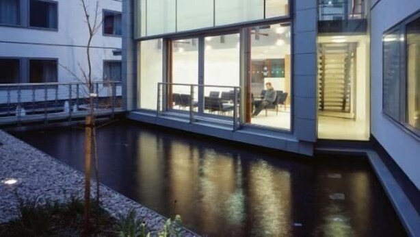 Ein modernes Gebäude mit großen Glasfenstern, einem Innenhof mit Wasserspiel und einer Person, die an einem Tisch sitzt.