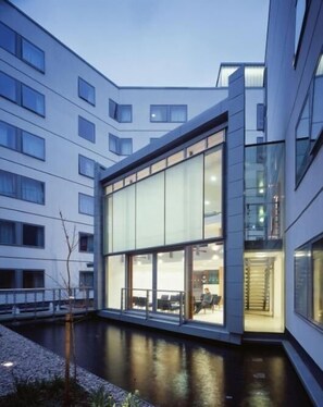 A modern building with large glass windows, a courtyard with a water feature, and a person sitting at a table. - Friendship House - Hostel (London)