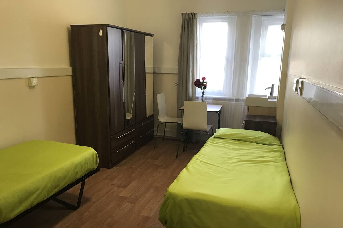 Una habitación con dos camas individuales, un armario de madera, un escritorio con silla y una ventana con cortinas.
