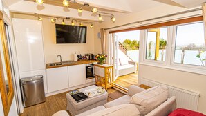 A modern kitchen with a built-in oven, a sink, and a flat-screen TV. There is a wooden dining table with chairs, a sofa, and a red throw blanket. - Sea View Holiday Flat - Covehurst Bay (Hastings)