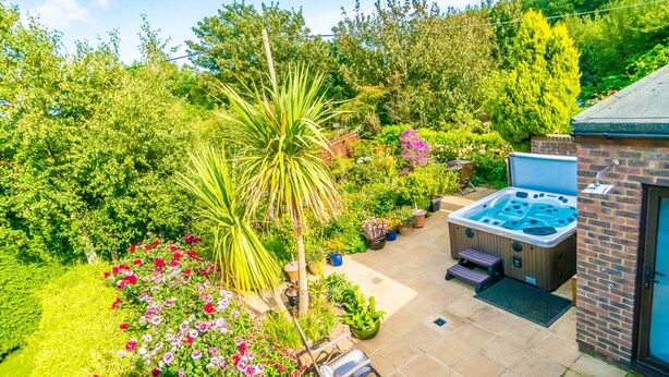 A patio with a hot tub, surrounded by lush greenery and flowering plants. - Covehurst Bay Holiday Cottages (Hastings)