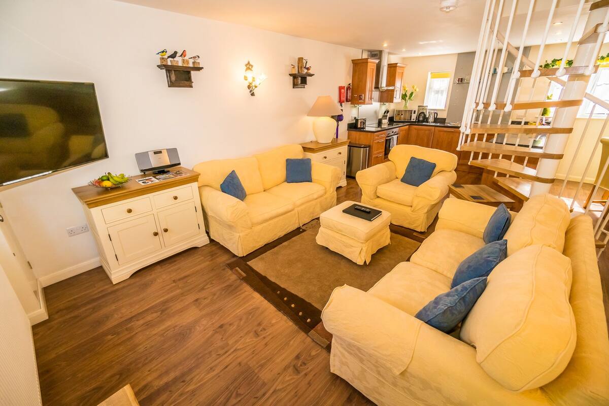 A living room with two yellow sofas, a wooden cabinet, a flat-screen TV, and a kitchen in the background.