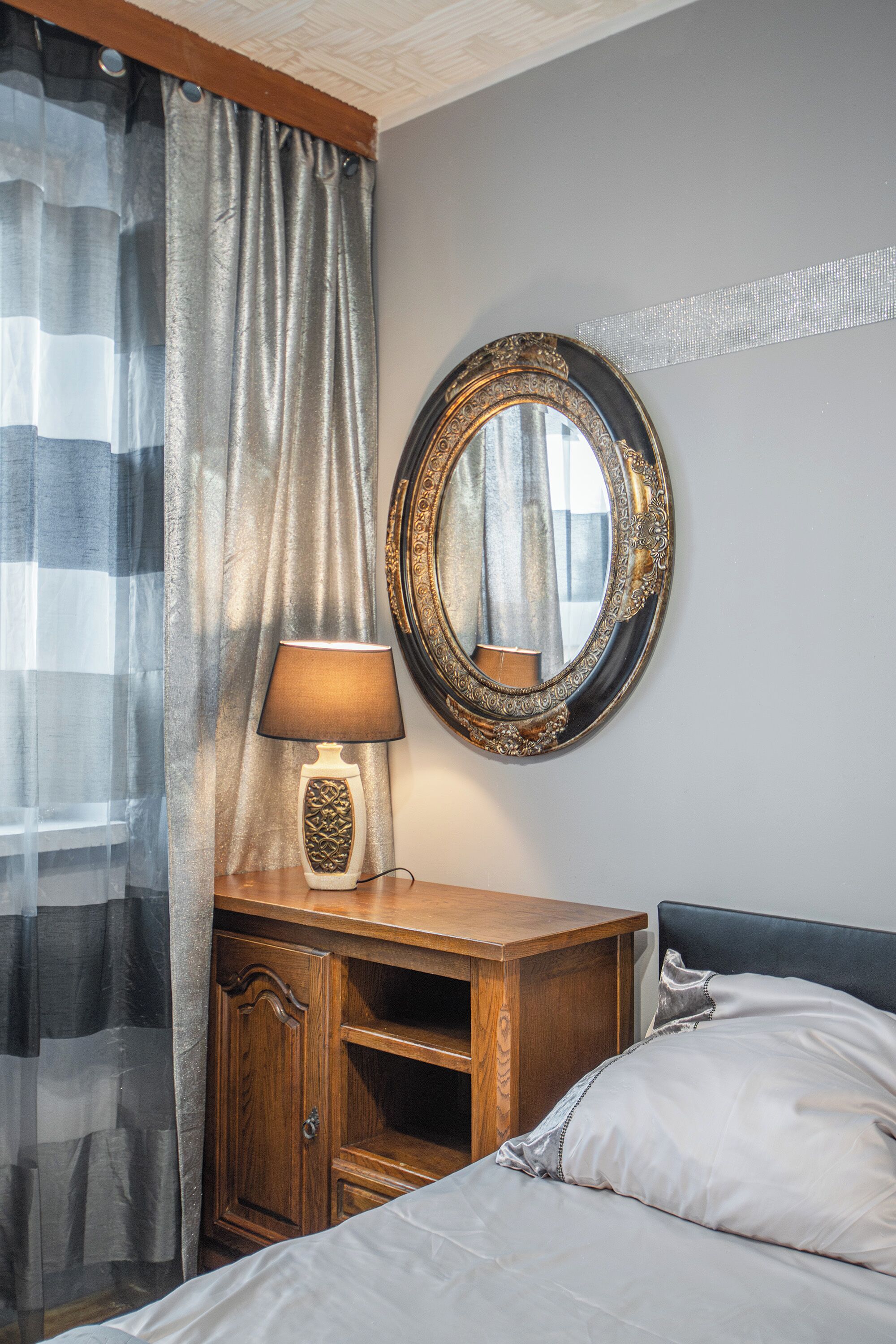 A bedroom with a bed, a wooden nightstand with a lamp, a mirror, and a window with curtains.