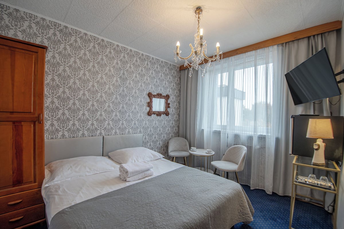 Une chambre d’hôtel avec un grand lit, une armoire en bois, un lustre, une télévision et une petite table avec des chaises.