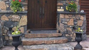 Entrada de pedra com porta de madeira, ladeada por luminárias e vasos de plantas.