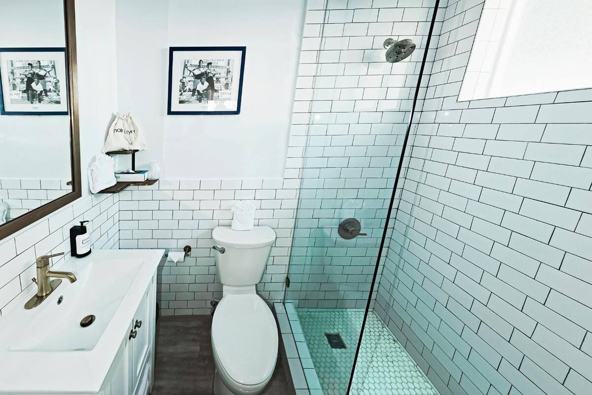 A bathroom with a white sink, toilet, and glass-enclosed shower. There are framed pictures on the wall and a small shelf with toiletries.