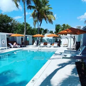 A pool area with lounge chairs and umbrellas, surrounded by palm trees and a clear sky.