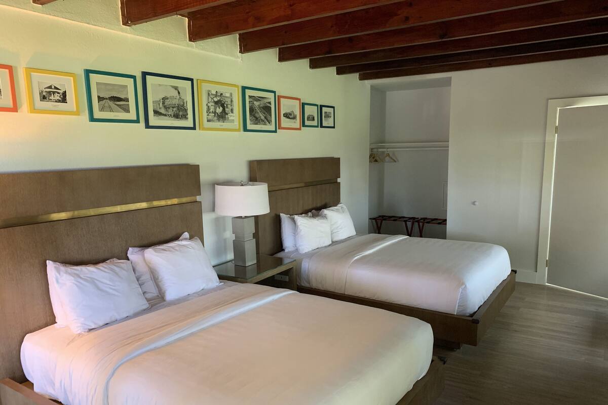 A hotel room with two beds, a wooden headboard, a bedside table with a lamp, and a wall with framed pictures.