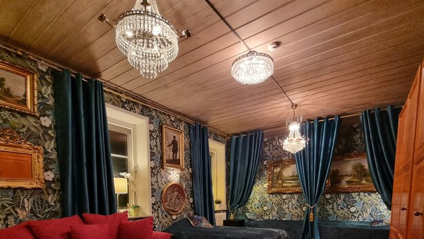 Une pièce avec un canapé rouge, un canapé noir, une table basse ronde et un grand tapis rouge à motifs. Deux lustres sont suspendus au plafond.
