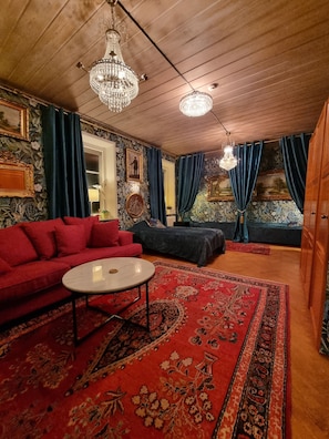 Een kamer met een rode bank, een zwarte bank, een ronde salontafel en een groot, roodgekleurd vloerkleed. Twee kroonluchters hangen aan het plafond.