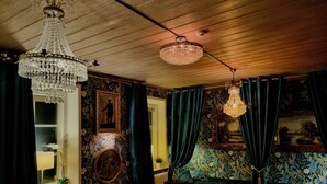 Une pièce avec un plafond en bois, un lustre, un lit, une fenêtre avec des rideaux et un mur recouvert d’un papier peint à motifs floraux.