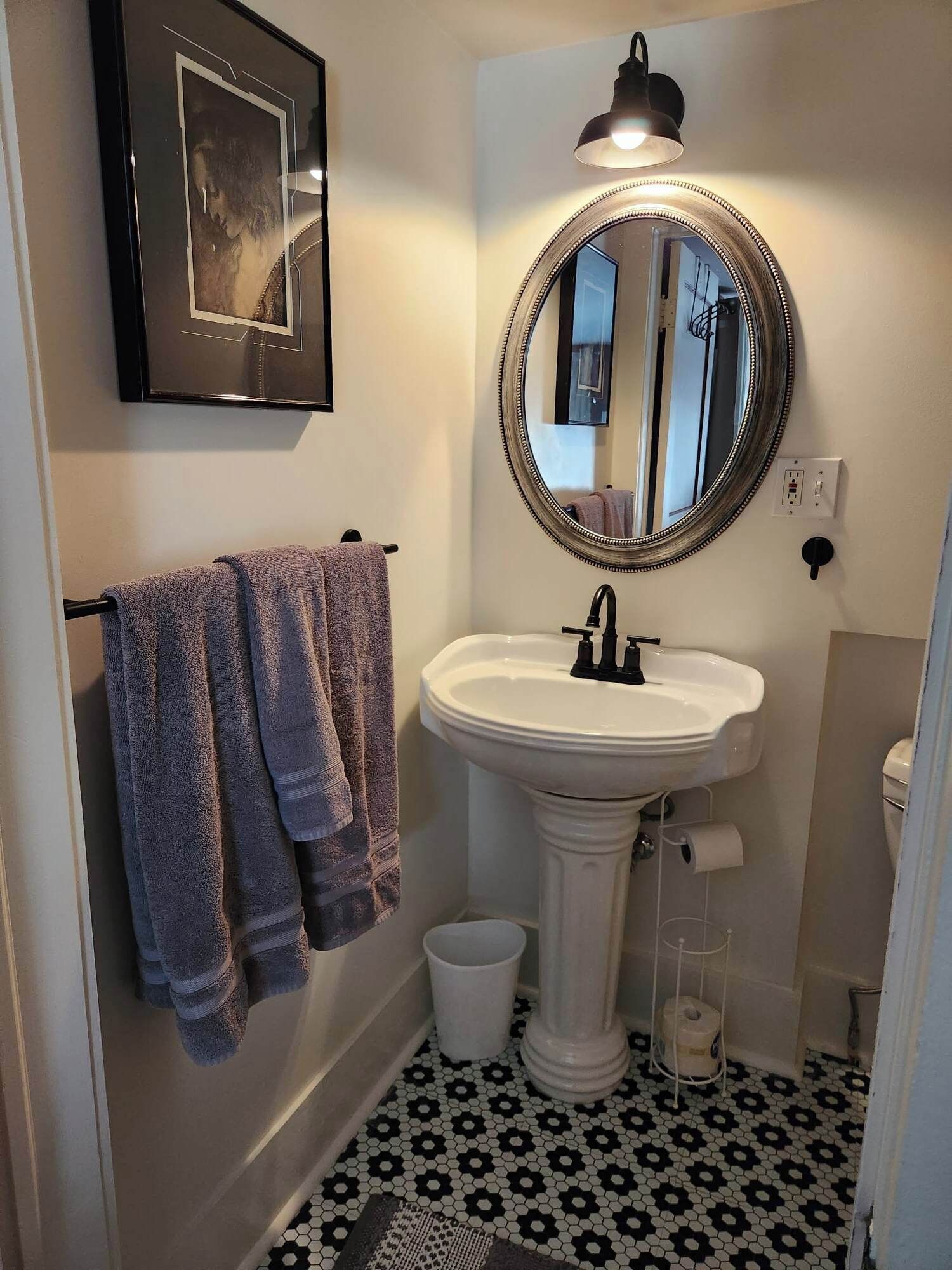 A bathroom with a pedestal sink, a round mirror with a decorative frame, a framed picture on the wall, and a black and white tiled floor.