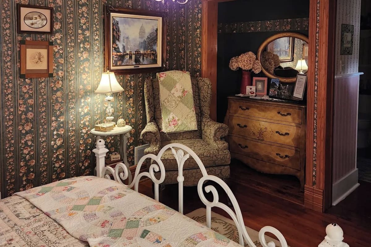 A bedroom with floral wallpaper, a bed with a quilt, a chair, a dresser, and a chandelier.