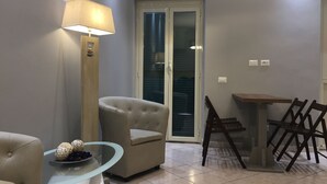 Ein Wohnzimmer mit einem beigefarbenen Sessel, einem Glastisch mit Dekoration, einer Lampe, einem Esstisch mit Stühlen und einer Schiebetür aus Glas.