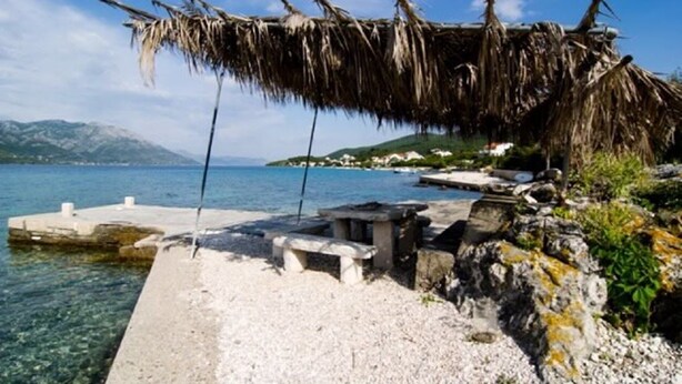 A coastal scene with a thatched structure, a concrete pier, and a rocky shoreline. - Apartmani MAK (Korcula)