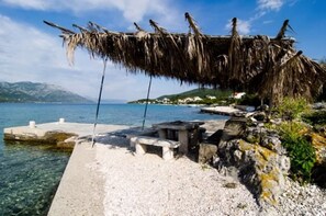 A coastal scene with a thatched structure, a concrete pier, and a rocky shoreline. - Apartmani MAK (Korcula)