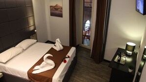 A hotel room with a bed, a nightstand, a TV, and a view of a cityscape through a window. - Dolce Vita Rooms & Breakfast (Naples)