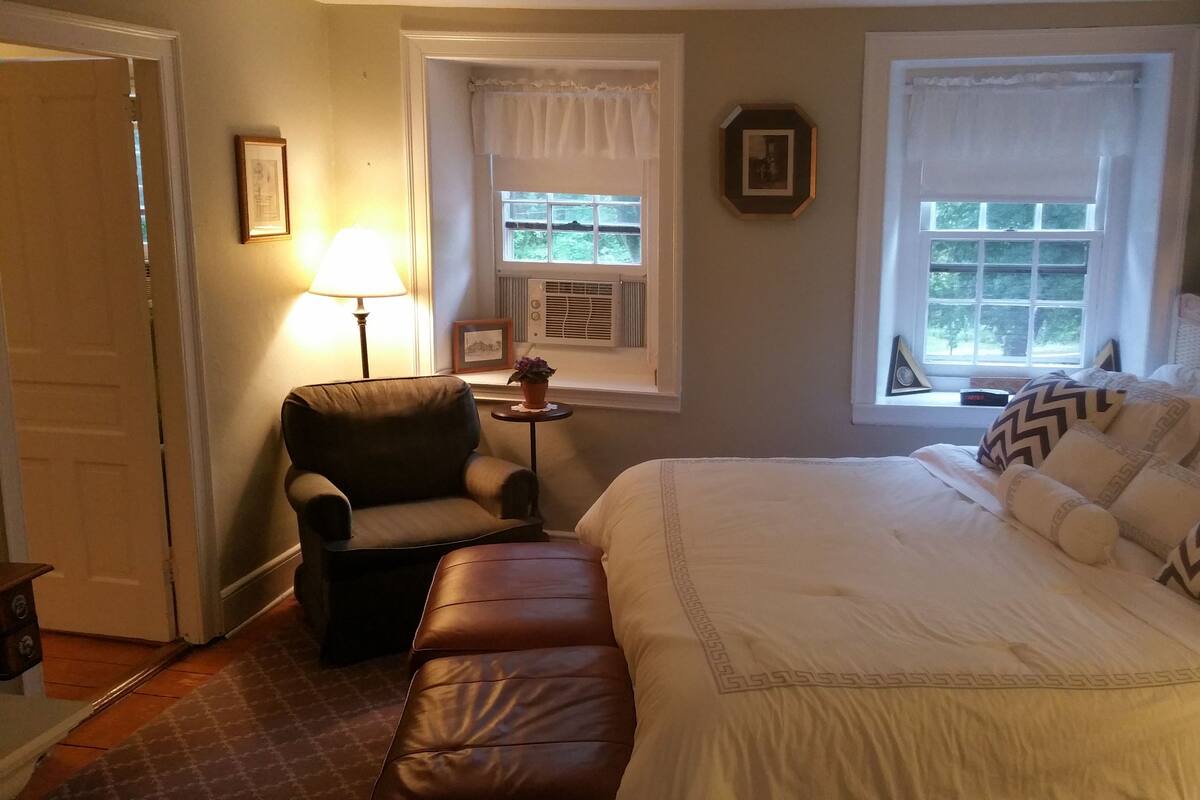 A bedroom with a large bed, a chair, a small table, and two windows with curtains.