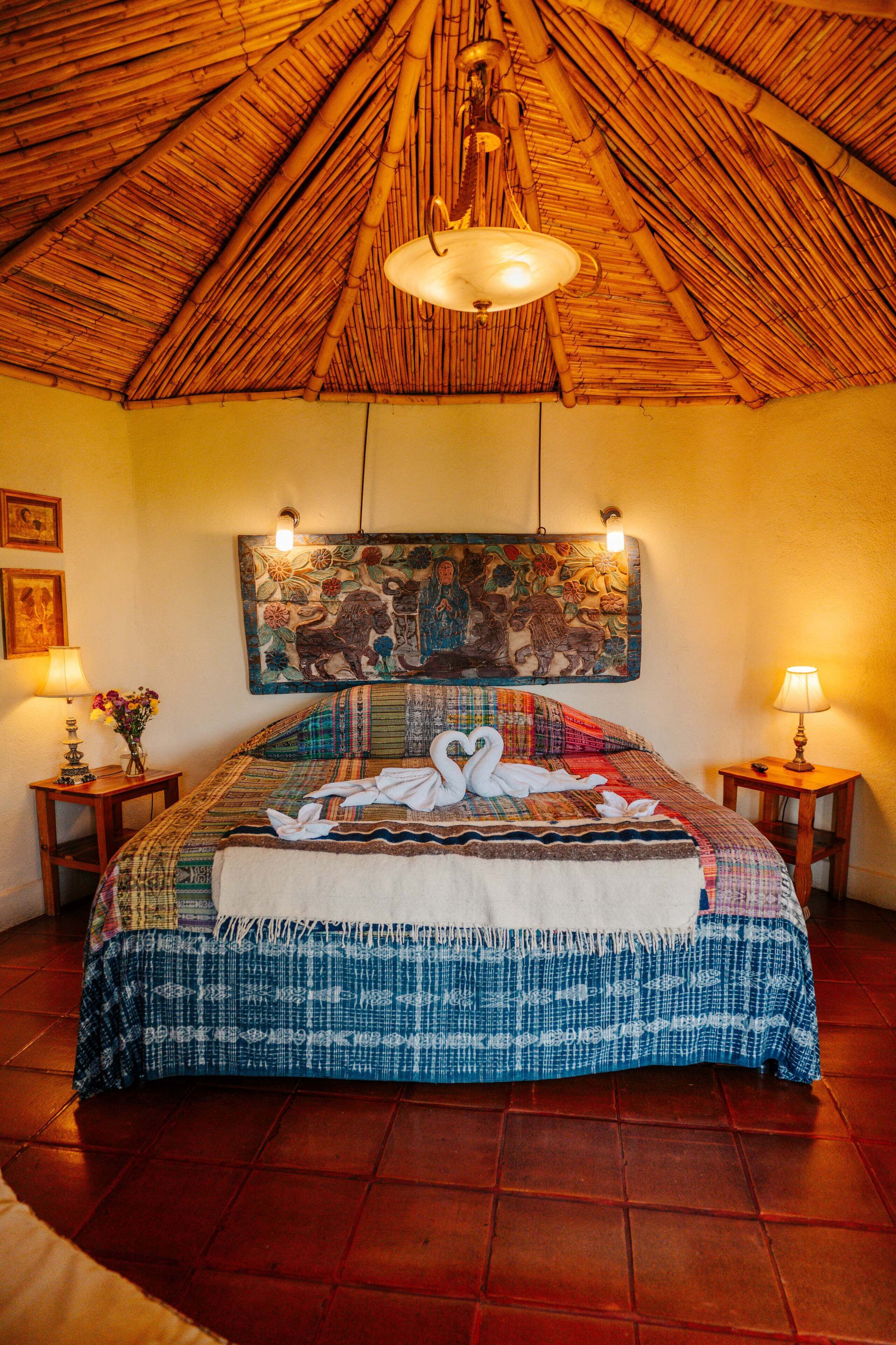 A room with a thatched roof, a bed with a colorful bedspread and a swan design, two bedside lamps, and a wall-mounted painting.