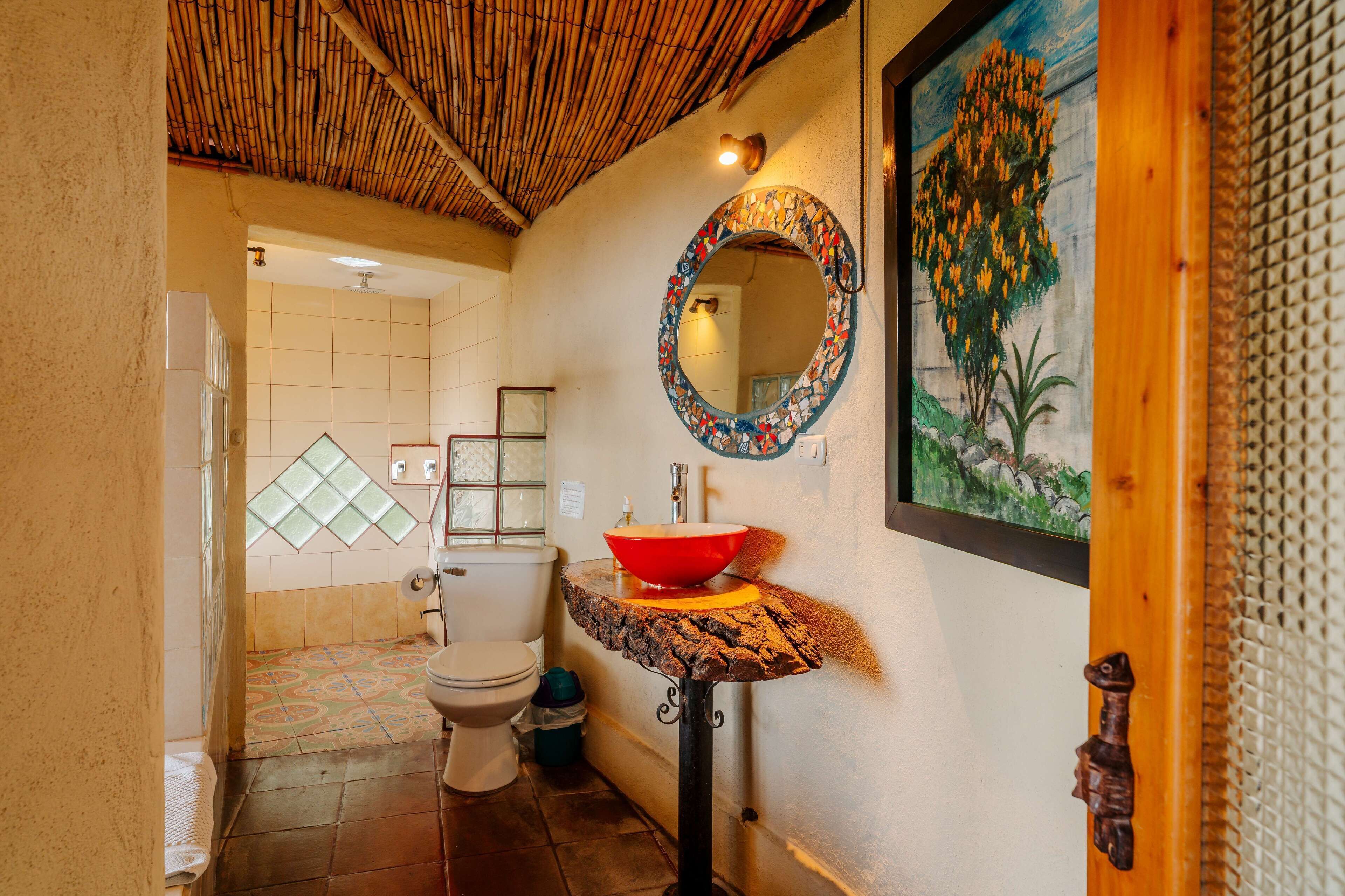 A bathroom with a bamboo ceiling, a round mirror with a colorful frame, a red basin, and a painting of a tree.