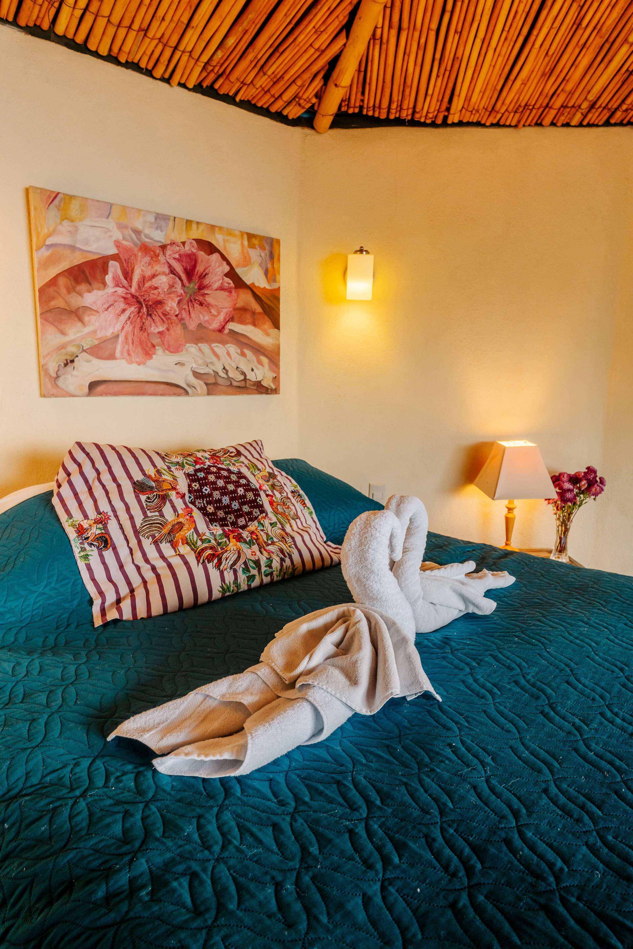 Habitación de hotel con edredón acolchado color turquesa, un cuadro floral en la pared, almohada a rayas, lámpara de noche y un florero con flores.