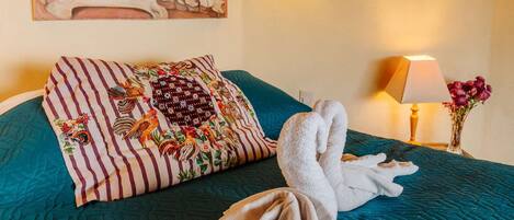Habitación de hotel con edredón acolchado color turquesa, un cuadro floral en la pared, almohada a rayas, lámpara de noche y un florero con flores.