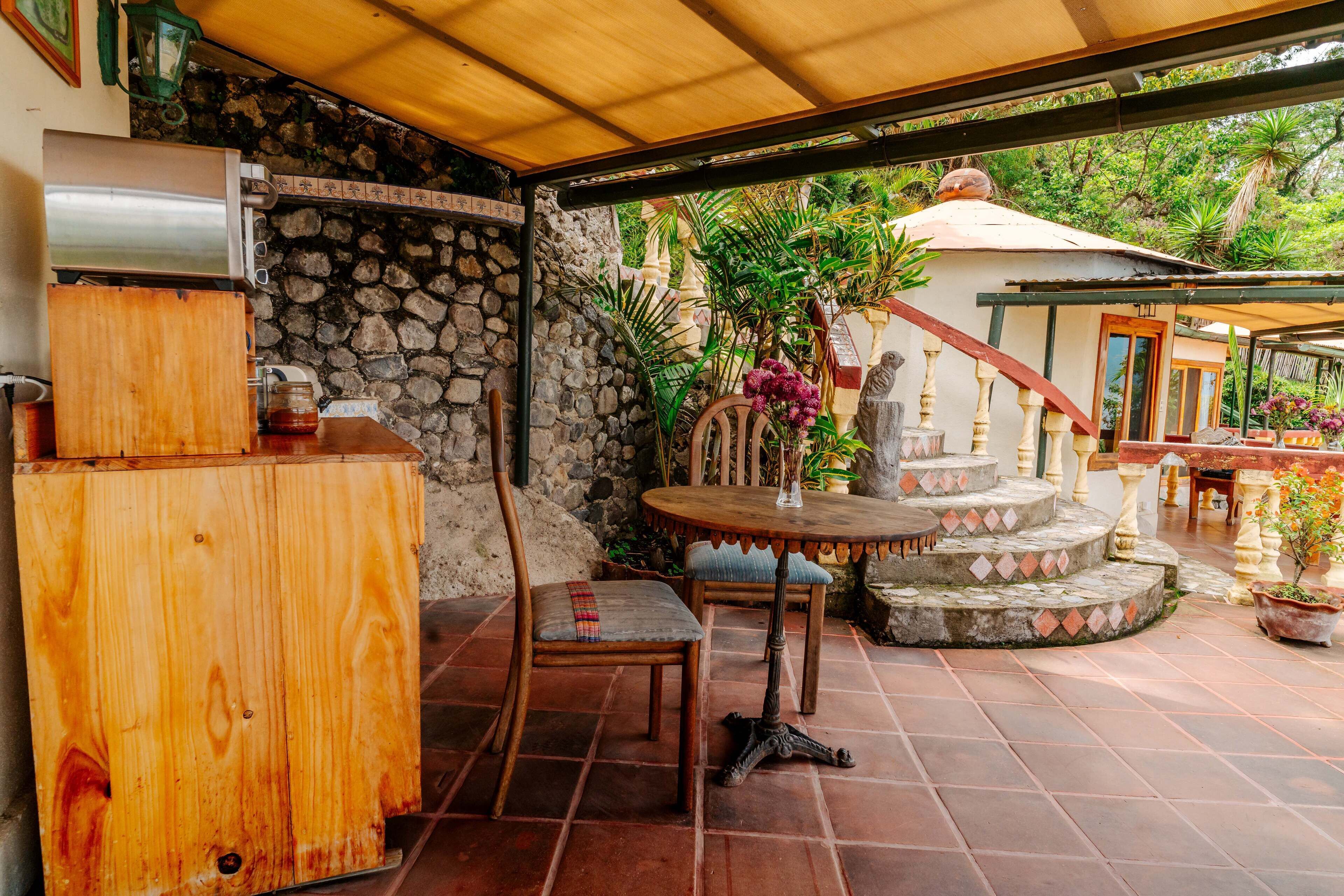 Une terrasse extérieure couverte, dotée d’un mur en pierre, d’une table en bois avec des chaises, et d’un escalier en pierre menant à une zone abritée.