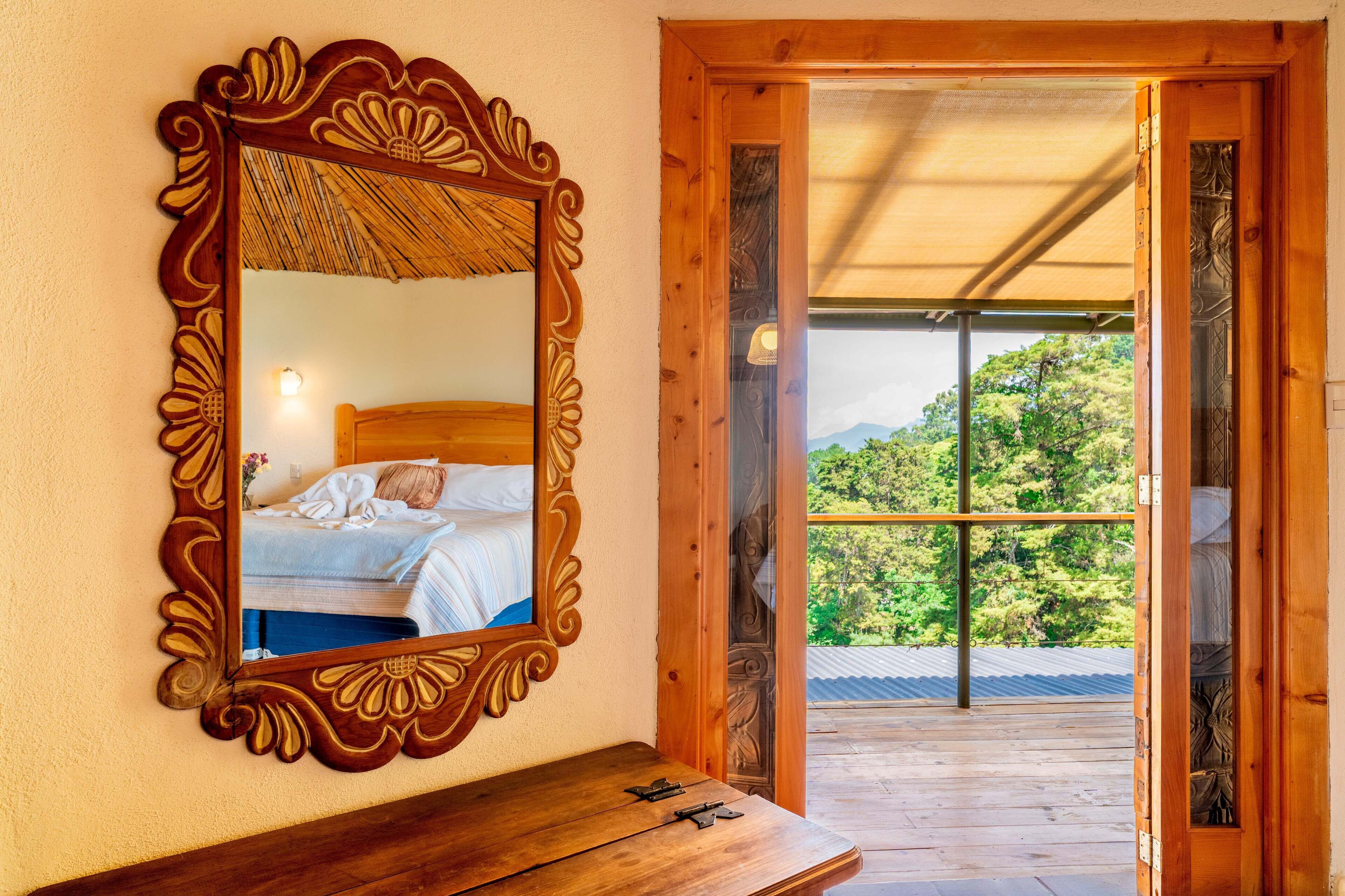 Une chambre à coucher dotée d’un grand miroir décoratif, d’une tête de lit en bois et offrant une vue sur un balcon avec un plancher en bois et de la verdure à l’extérieur.