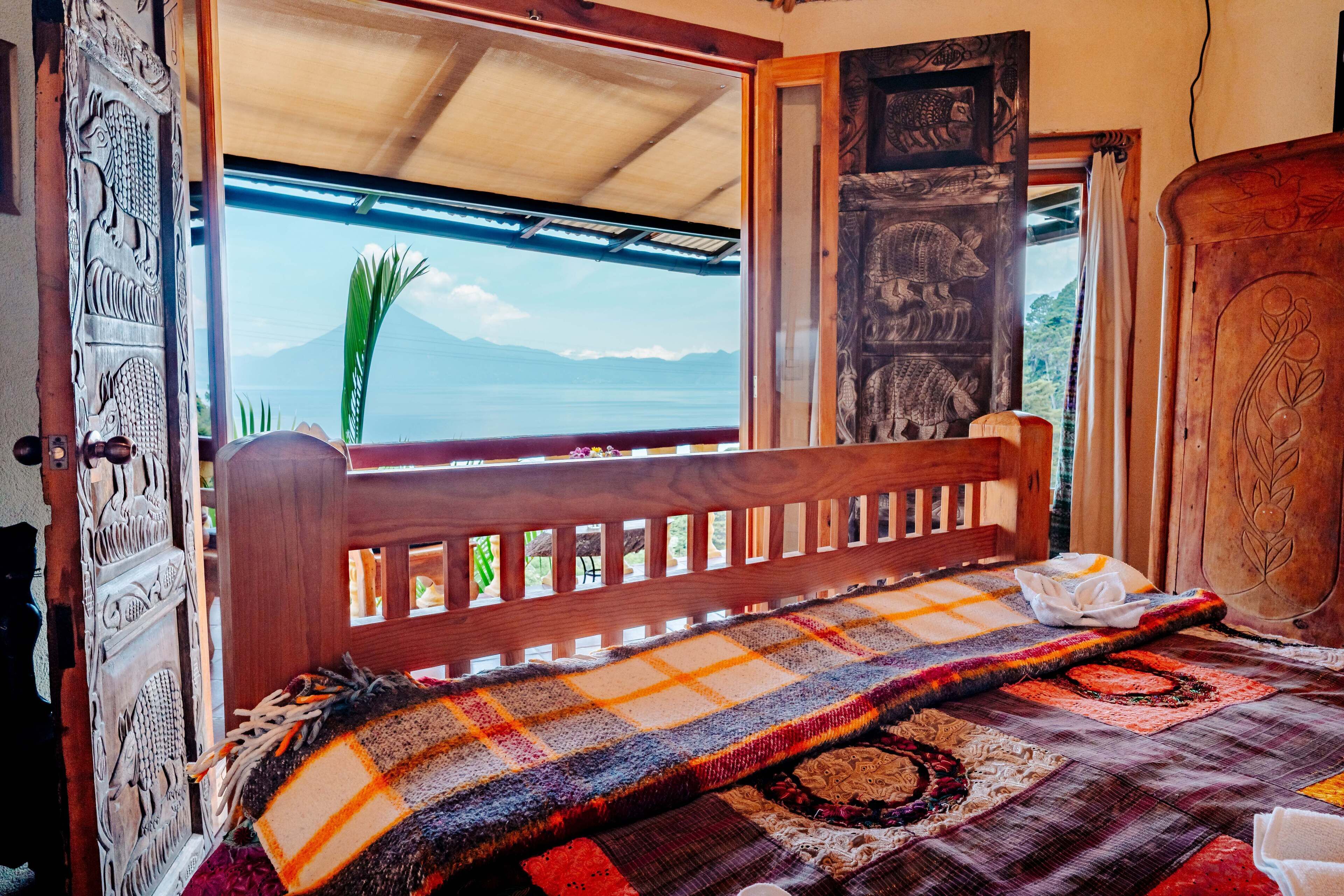 A bed with a colorful blanket, a wooden headboard, and a view of the sea through a window.