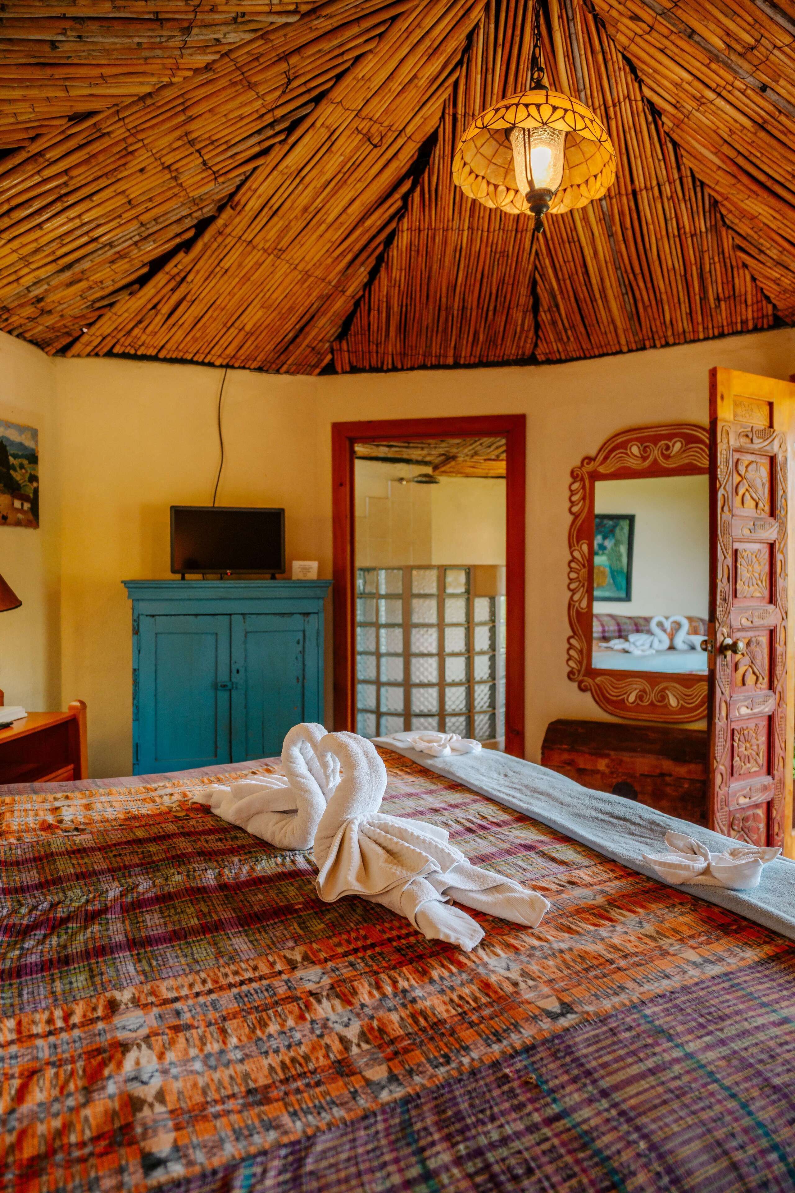 Una habitación con techo de paja, una cama con una manta de colores, un televisor sobre un mueble y un espejo con marco decorativo.