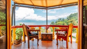 A balcony with two wooden chairs, a small table with a vase, and a view of a lake and mountains.