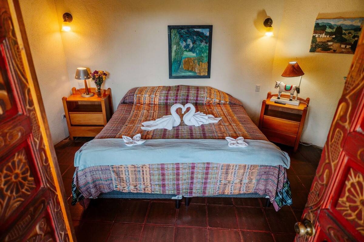 A neatly made bed with a colorful bedspread and two swan-shaped towels. There are two bedside tables with lamps and a painting on the wall.
