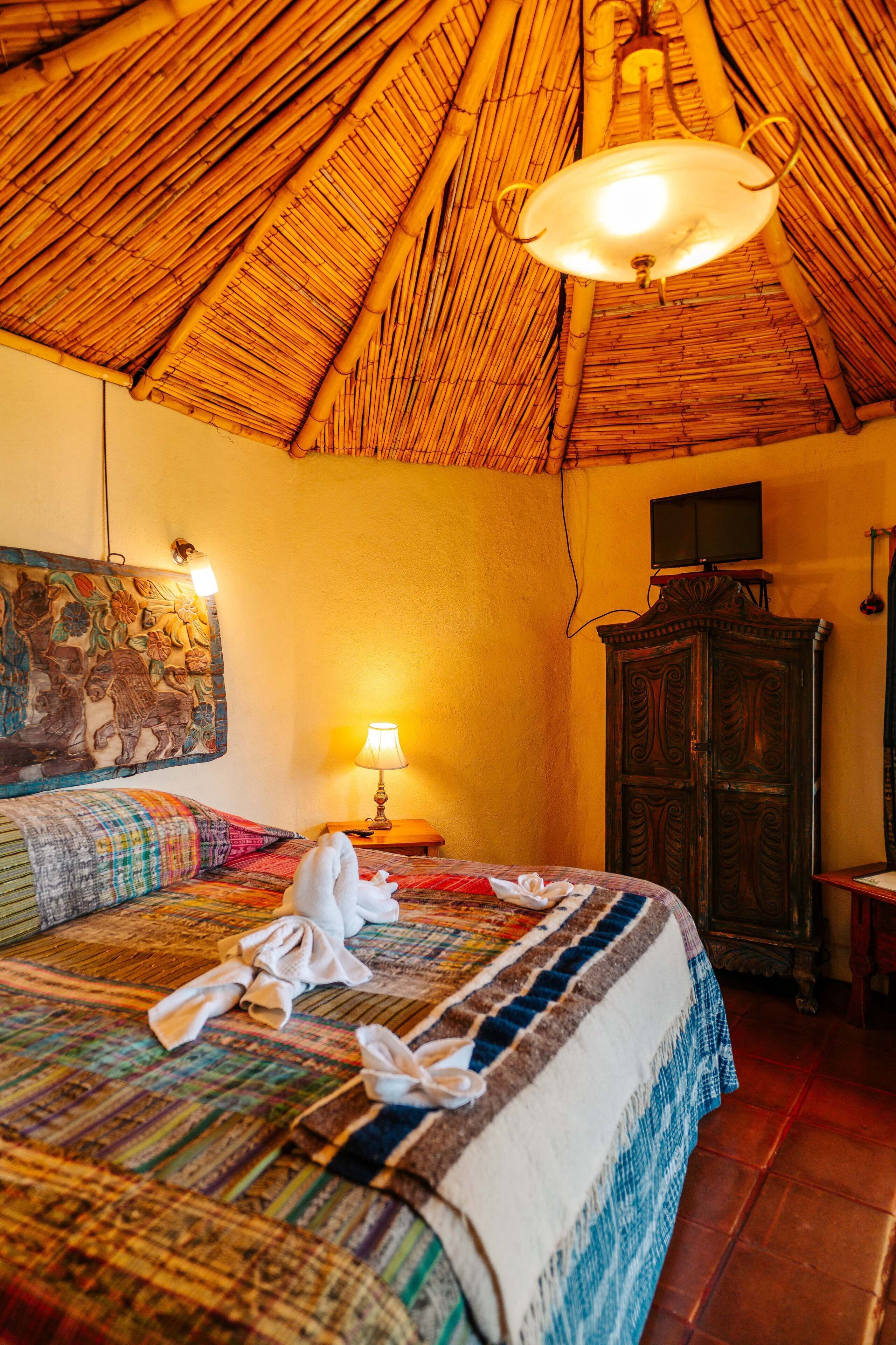 Una habitación con techo de paja, una cama con colcha de colores, un armario de madera y un televisor montado en la pared.