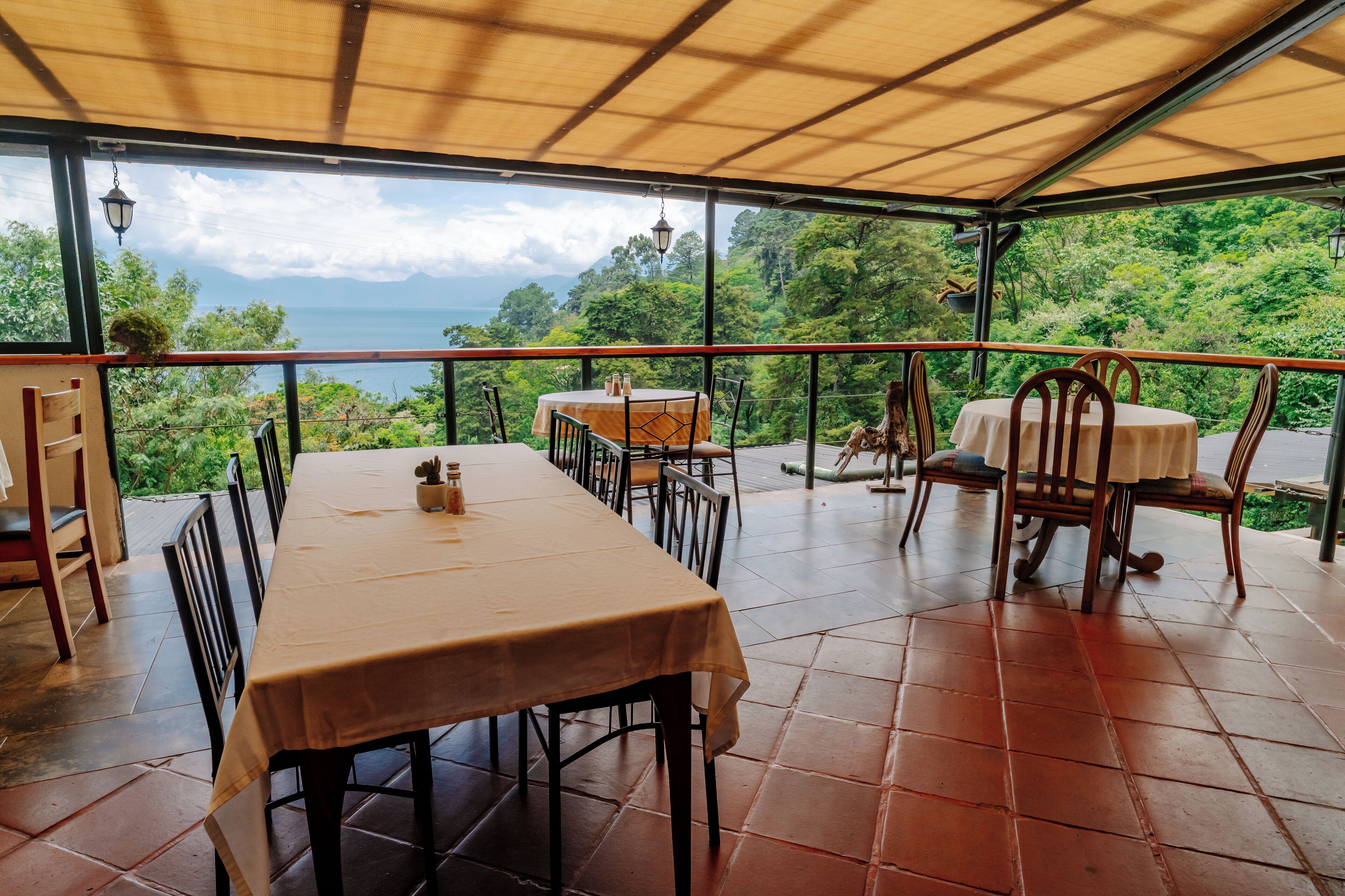 A covered terrace with a table and chairs, overlooking a lush landscape and the sea.