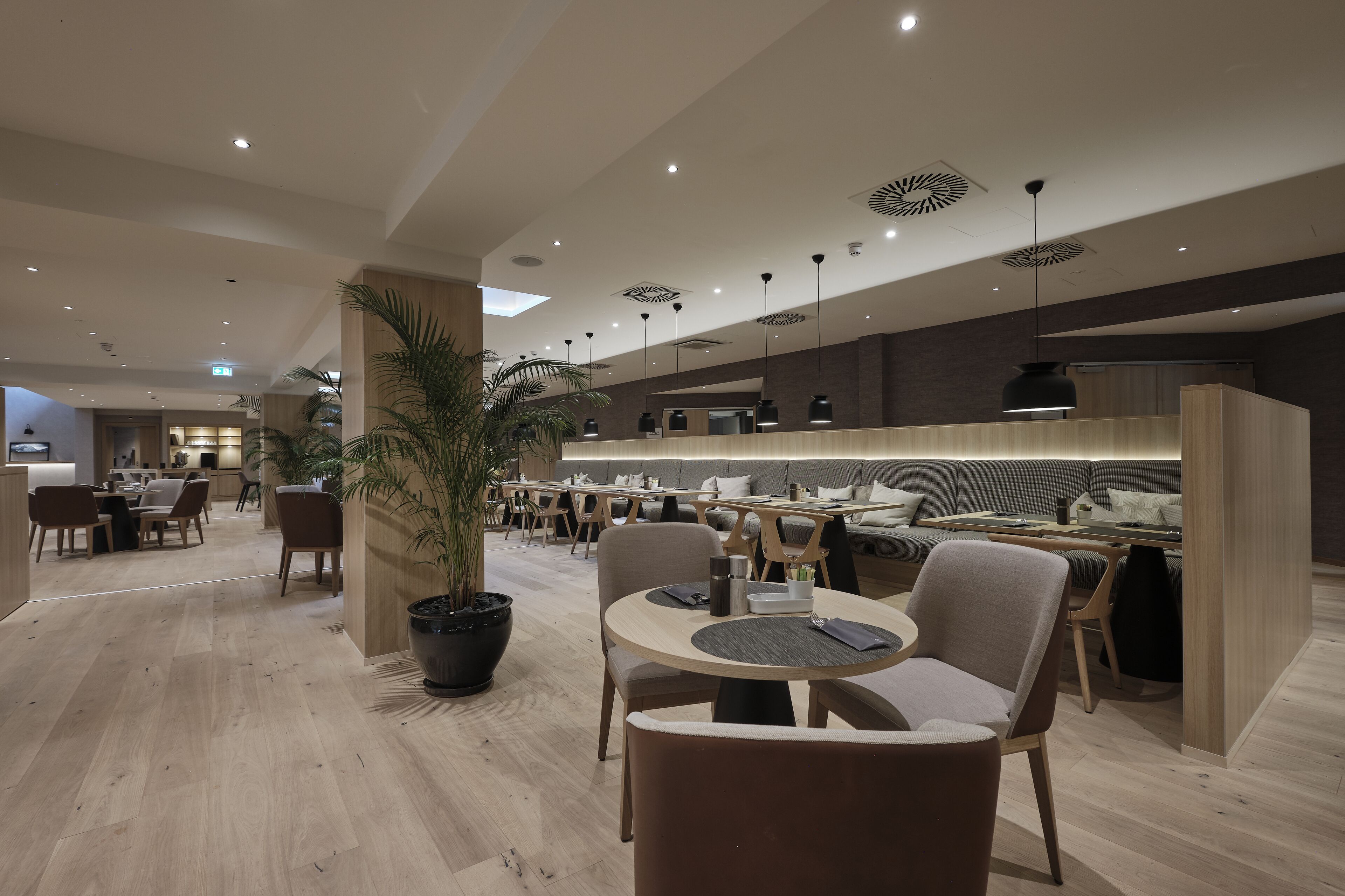 A modern restaurant with wooden flooring, round tables, and chairs. There are potted plants and pendant lights.