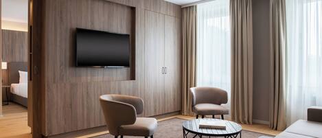 Una habitación de hotel moderna con una gran televisión de pantalla plana montada en la pared, dos sillones, una mesa de centro redonda y un amplio ventanal con cortinas.
