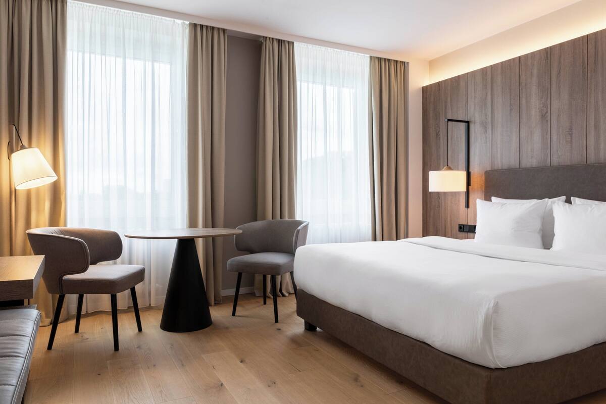 Una habitación de hotel con una cama grande, dos sillas, una mesita y una lámpara.